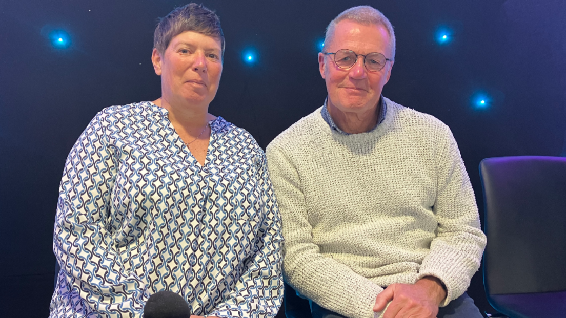 A woman with short dark hair, and a man with short hair and round glasses sit side-by-side looking at the camera