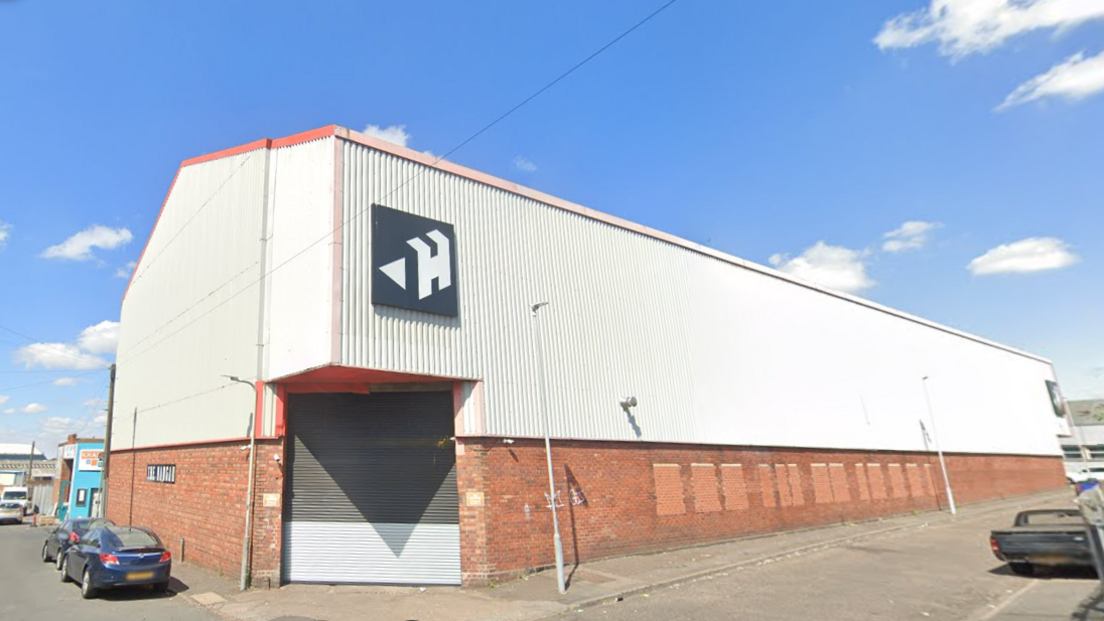 A large white warehouse on a street corner on a sunny day. The bottom section of the building is brick, and the top half is white and corrugated. There is a black logo with a capital "H" and a small triangle on the front.