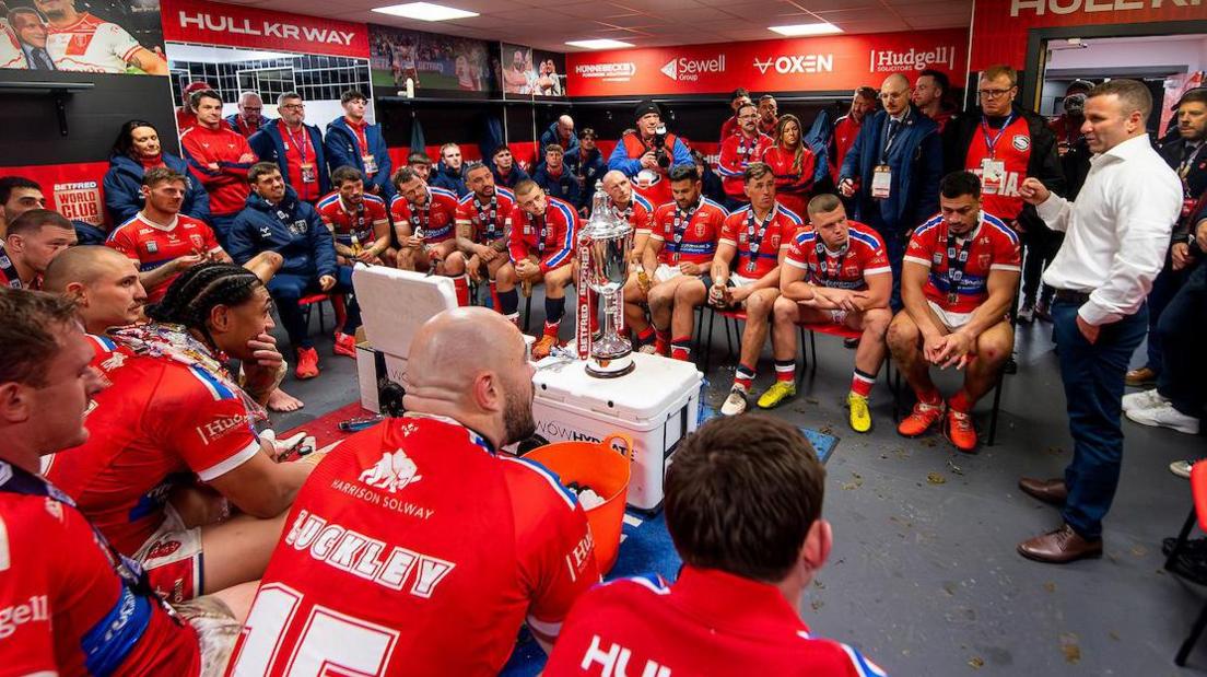 Hull Kingston Rovers coach Willie Peters, dressed in white shirt, black trousers and brown shoes, speaks to his Hull KR players, sitting around him in the dressing room, with the World Club Challenge trophy sitting on a white kit box in the centre.
