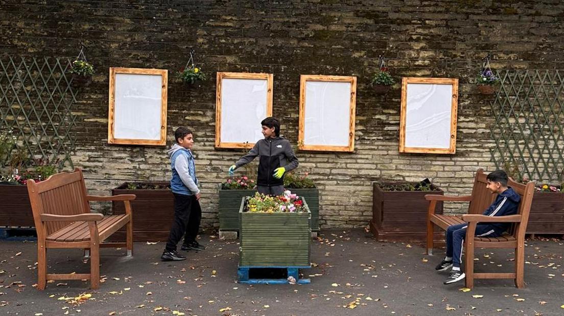 Three young boys relax and chat in a small community garden. There are two benches and a number of planter boxes with colourful flowers in them.