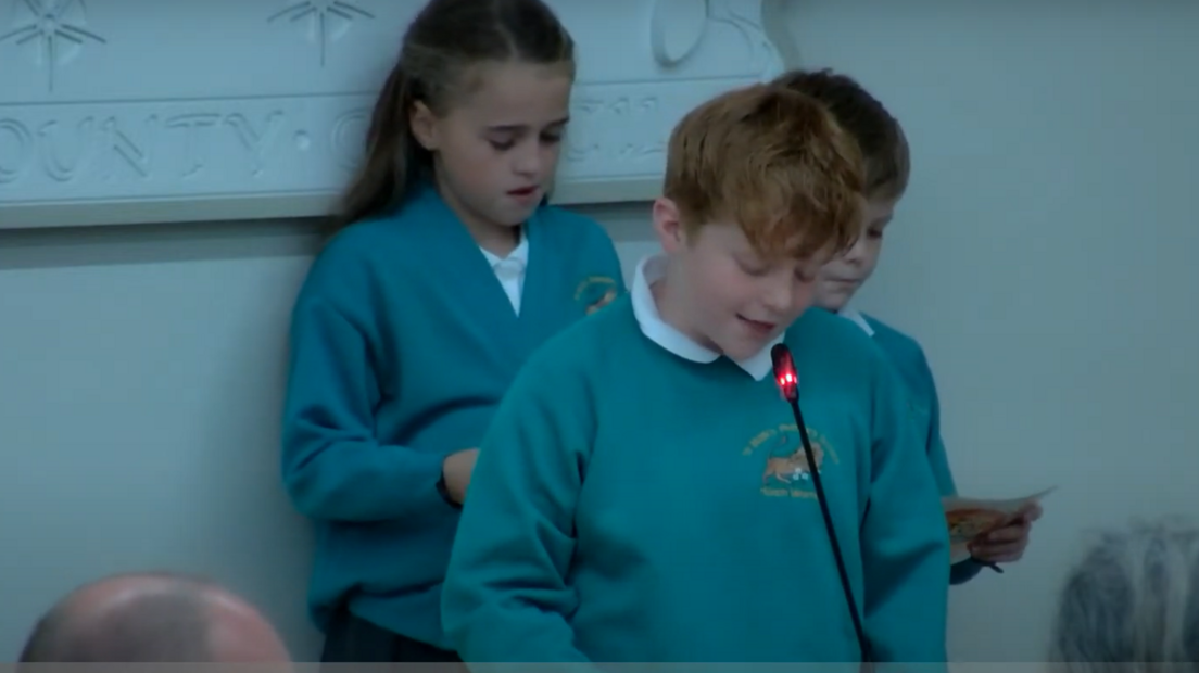 Two boys and a girl standing at a council meeting. They are wearing green school uniform tops and one boy is speaking into a microphone