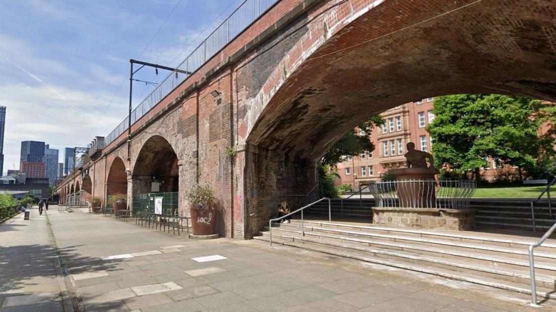 Red bricked archways loom over a street on a sunny day with tower blocks seen on the skyline in the distance. 