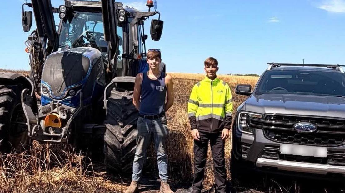 Two young men, Cameron and Jay, stand in a field. A tractor and pick up truck are next to them. The crops behind them are brown and burnt.