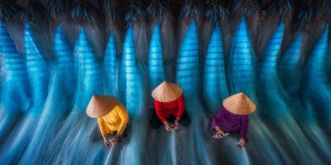 three figures, their faces hidden by hats, shown working with swathes of blue threads