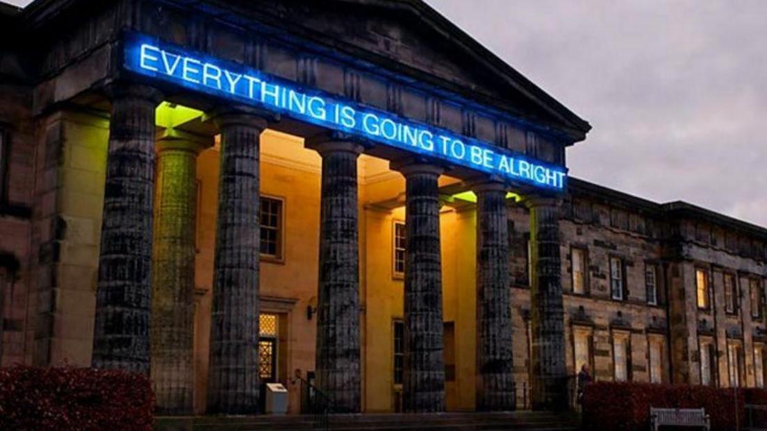 A blue neon sign reading "everything is going to be alright" is installed on a large building with six pillars.