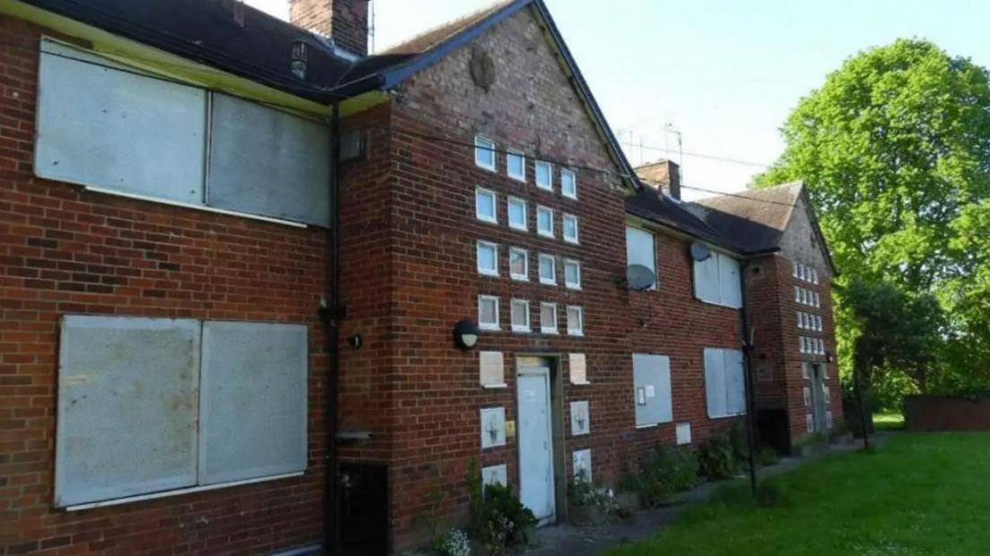 The exterior of Henson Villas in Pearson Park. It is a red brick building with multiple large windows boarded up, as well as multiple small windows. There is grass in front and green trees can be seen in the background.