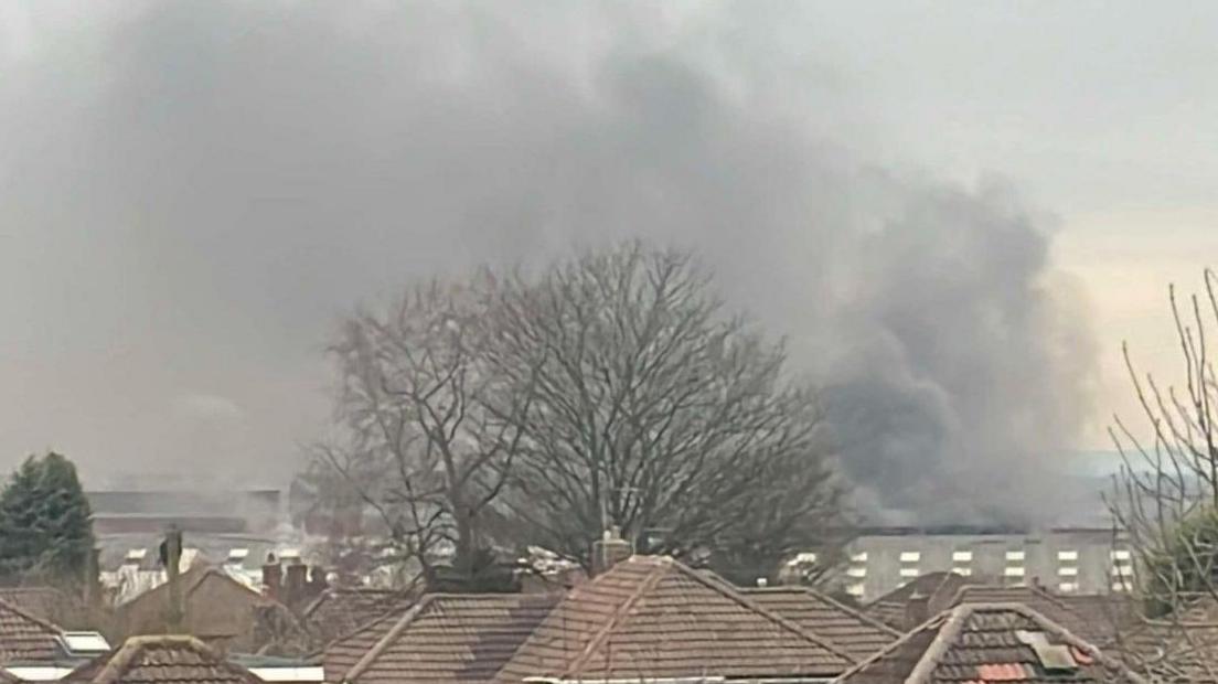 Smoke going high into the air in the distance. Rooftops and some trees are in the foreground.