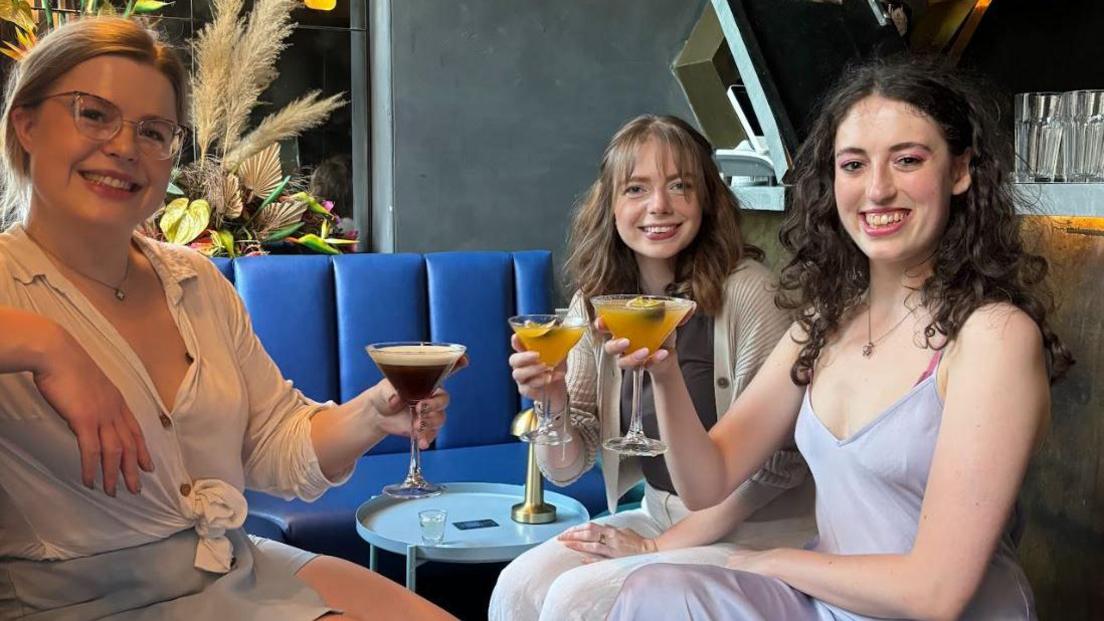 Three young women, including Olivia Bevan, sitting around a table drinking cocktails on a blue velvet bench smiling. Olivia is blonde, wearing glasses with a white top and silver skirt, she's holding an espresso martini. The other two young girls are brunettes. One is wearing a strappy top  and the other a cardigan. They're holding fruity orange cocktails. 