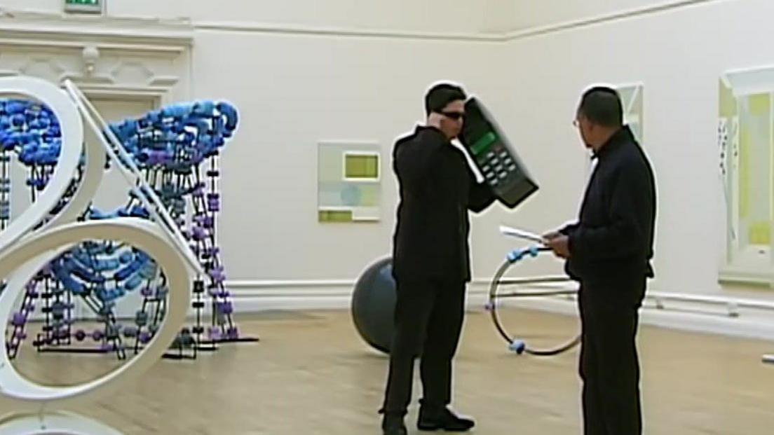 Dom Joly is stood inside a museum with art all around. He is wearing black sunglasses, and holding a giant telephone and shouting "hello?". Stood next to him is a man looking at him.
