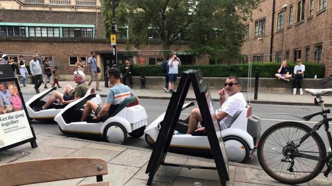 Three men are riding their C5s along a street in Cambridge. A man can be seen taking a photograph of them from the other side of the street. People are sitting on a college wall and others are walking along the pavement. There are advertising boards on the street in front of the C5s