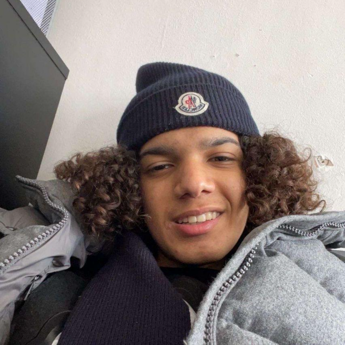 Mohamed Abdi is seen with curly hair wearing a dark beanie and a grey padded jacket smiles while reclining indoors against a light-coloured wall.