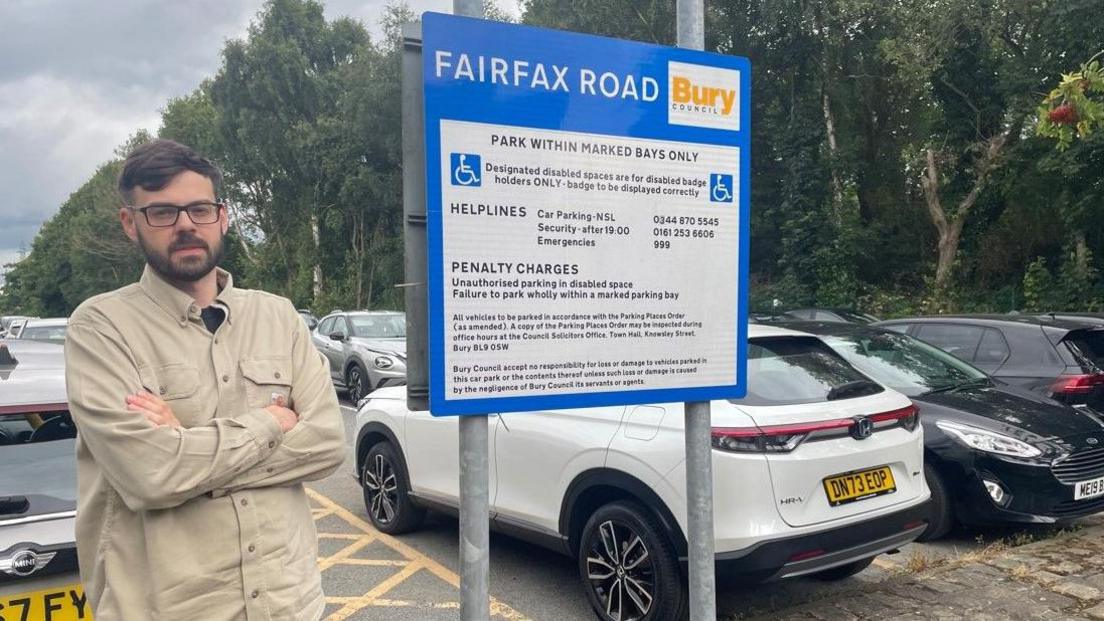 Isaac Jones stands with his arms folded in front of a car park information board at the Fairfax Road car park. There are a number of cars parked behind him, and a line of trees.