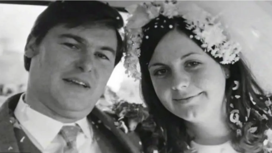 Black and white wedding photo of a man in a white collar suit and a woman wearing a flower veil. Both have confetti in their hair and on their shoulders 