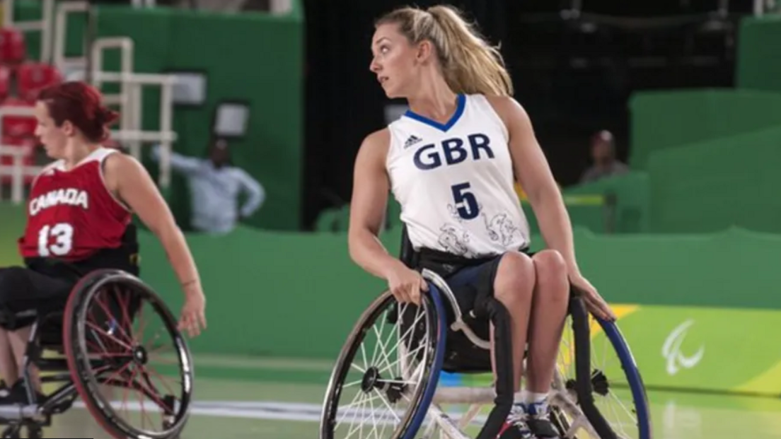 A woman in a wheelchair wearing a white and blue vest with GBR on it, looking over her left shoulder. Behind her is another woman in a wheelchair wearing a red vest that says Canada.