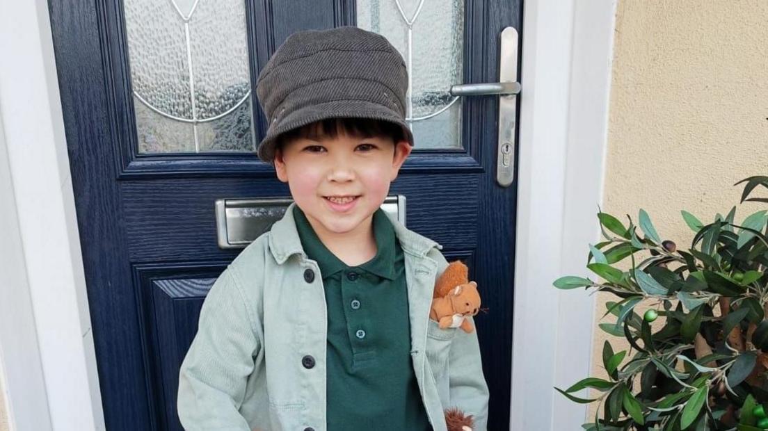 Joey is standing in front of a blue door and is wearing a green T-shirt and lighter green over shirt, with a toy squirrel in the top pocket. He is smiling and is wearing a grey hat. 