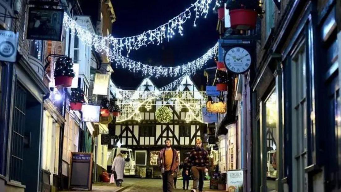 Dangling white lights pictured across the top of The Strait in Lincoln, with yellow lights in the distance which have bell shapes. A few people are seen walking up and down The Strait and it is evening. Independent shops can be seen either side of the street with a Tudor building in the distance.