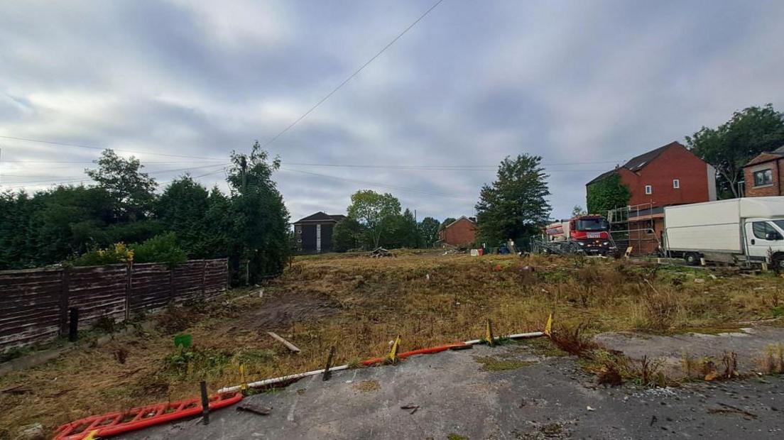 A derelict area of neglected land filled with weeds, concrete and knocked over fences.