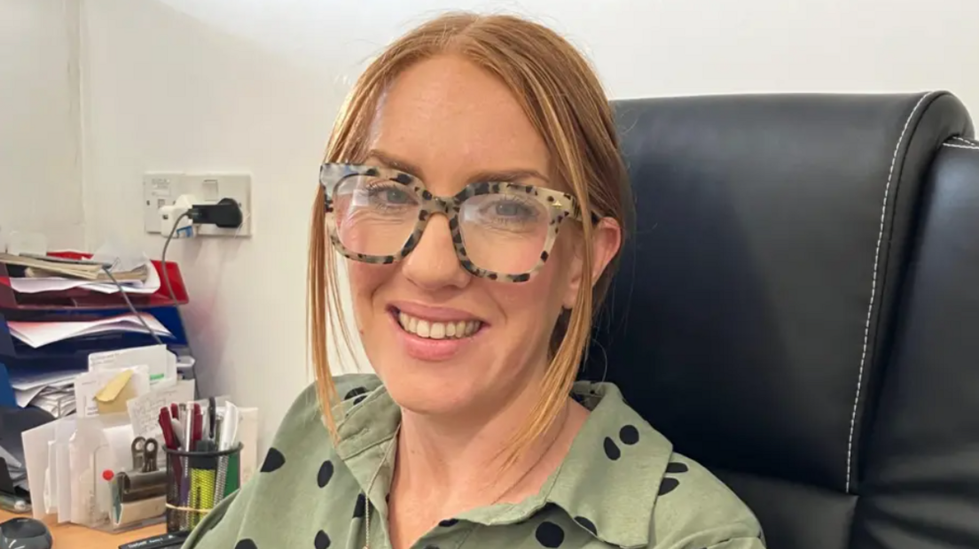 Stacey Lock sitting at her desk, smiling at the camera. She is wearing leopard-skin print glasses and a green top with black dots on it. 