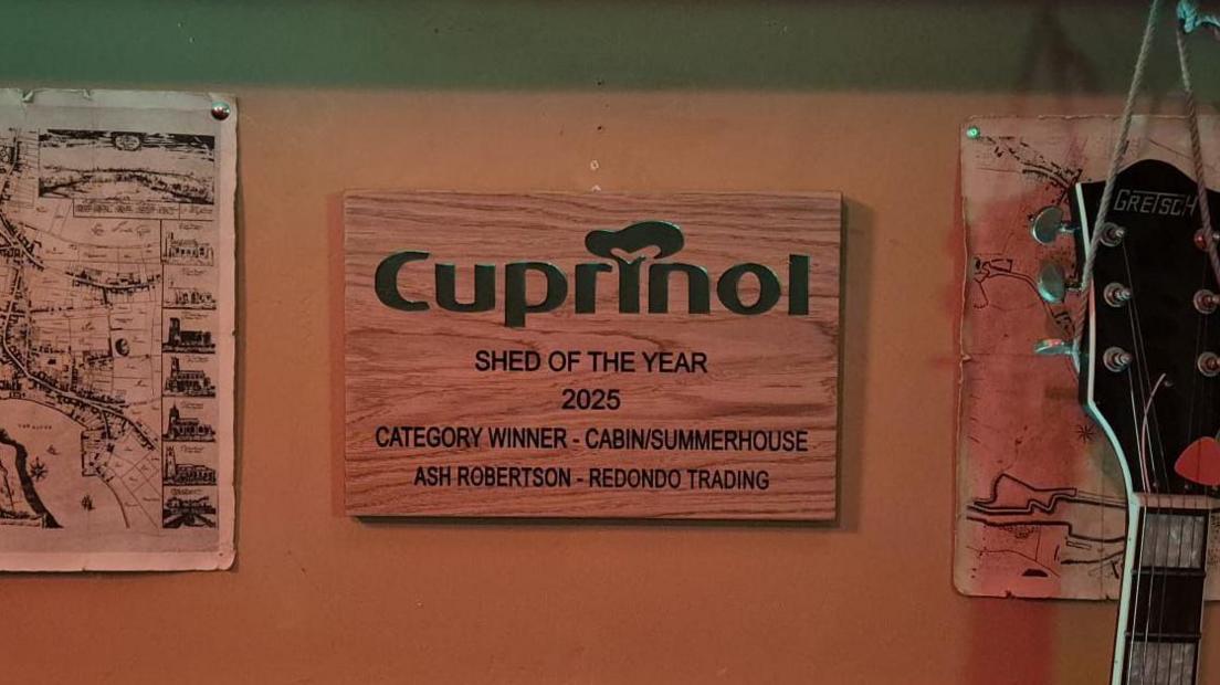 A wooden plaque reads: "Cuprinol. Shed of the Year 2025. Category Winner - Cabin/Summerhouse. Ash Robertson. Redondo Trading." 