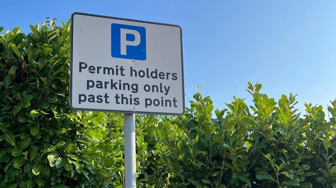 A square shaped parking sign on a pole, showing  a blue and white logo and the words 'Permit holders parking only past this point'