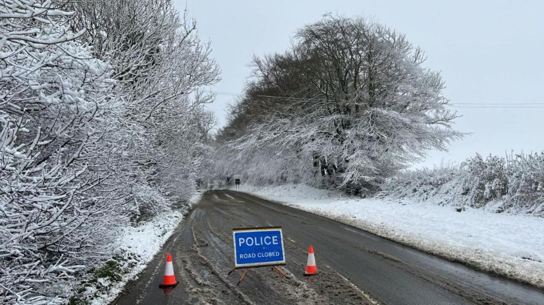 A road that is going though snow covered trees. There is a hedge on the right also covered in snow. The road has brown slush built up on either side and down the middle and in the foreground there is a blue 'Police Road Closed' sign and two bright orange traffic cones.  