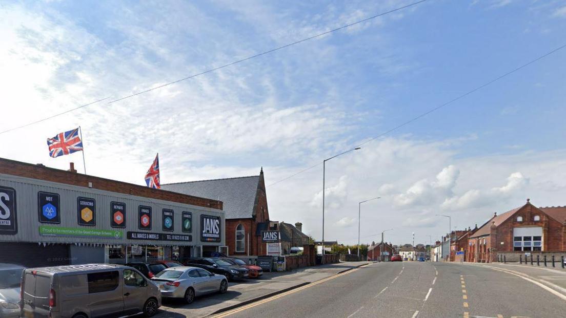 Streetview image of Mansfield Road, Sutton in Ashfield, showing a industrial unit with cars outside and, a short distance further along, a red brick primary school building.