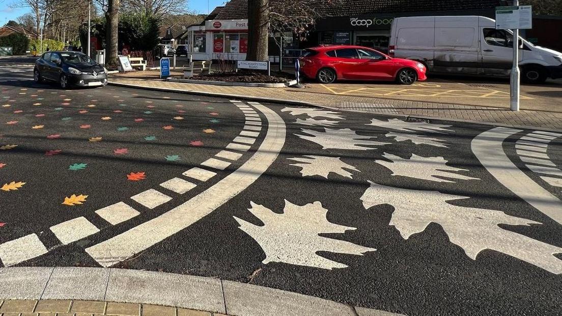 A photo taken from the pavement showing one of the roundabouts, with the white leaf crossing on the right and the rainbow leaf roundabout on the left. It's a sunny day and the sky is blue.