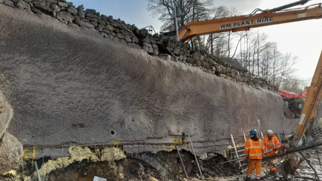 A crane reaching over a stone wall to a much deeper drop on the far side. Two people in orange high-visibility clothing are working at the end of the crane arm. The wall sits on top of a concrete surface, with heavy wire mesh visible at the bottom in front of old bricked arches. 