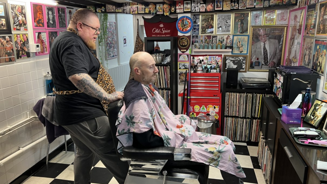 Wil Hodgson standing behind a barber's chair in which a client is sitting with a floral gown over him. The parlour walls in the background are covered in photographs, posters and memorabilia and there are shelves with vinyl records