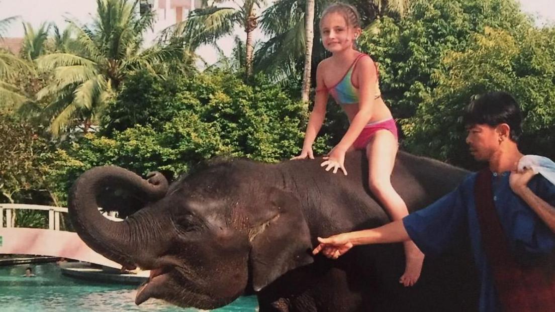 Amber Mason, sitting on a elephant, she is wearing a bikini, that is multi-coloured. Her bottoms are pink, there is a man to the right, holding onto the elephant, wearing a blue top. He ha short dark hair. Amber has her hair braided back. 