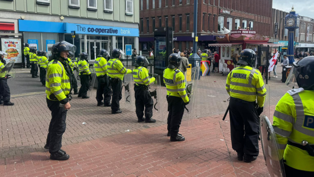 A line of officers in helmets and with shields stand in the centre of Hanley