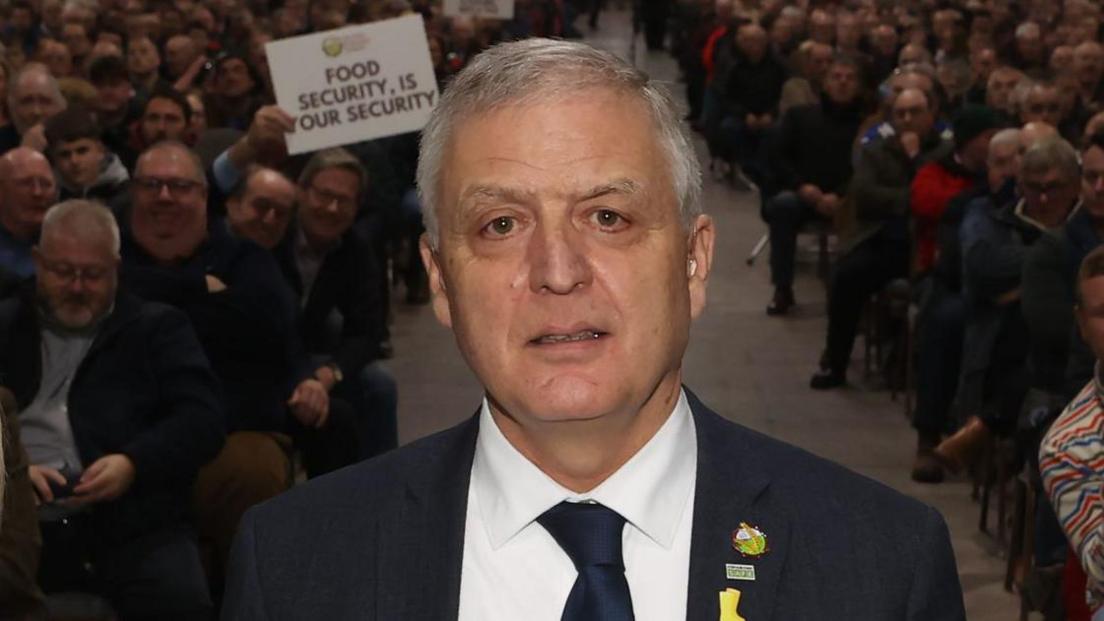 William Irvine - a man with short grey hair wears a dark suit jacket with some pins on the lapel, with a white collared shirt and a dark tie. He is looking at the camera. Behind him are people sat down in chairs, with one holding a sign saying 'food security is your security'.