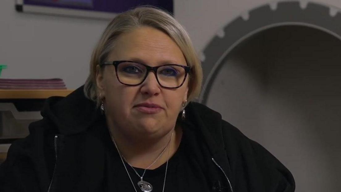 Childminder Victoria Cook. She has short, blonde hair, glasses and is wearing a black zip-up jacket and has silver necklaces on. In the background is a mirror.