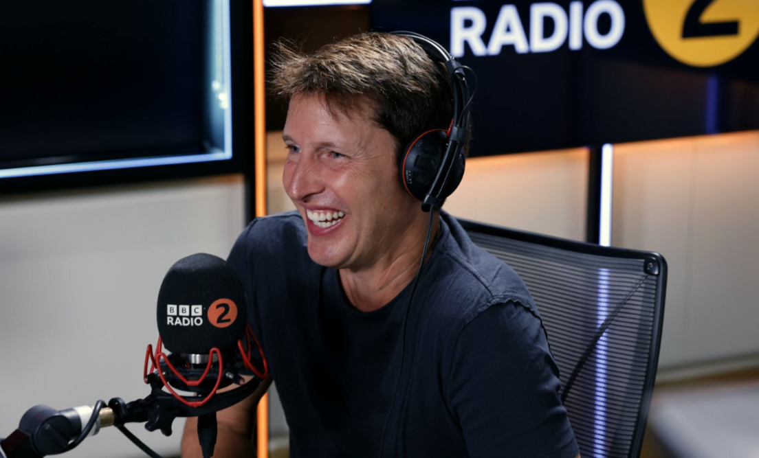 James Blunt laughing during radio interview with a large mic in front of him