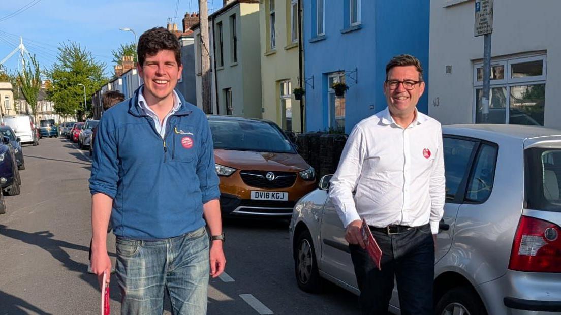 Mae'r llun yn dangos Huw Thomas ac Andy Burnham allan yn ymgyrchu ar ddiwrnod braf. Mae'r ddau'n gwenu.