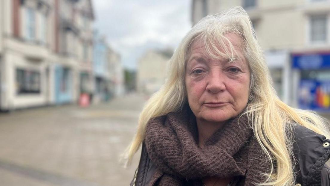 Penny Lloyd wearing a cowel-neck brown sweater and a brown coat and standing in the Triangle in Teignmouth with the street in a blurred background.