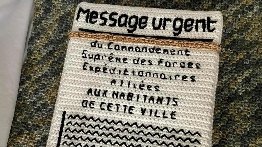 A knitted white papered message in French which says 'Message Urgent' and warns residents to leave a town