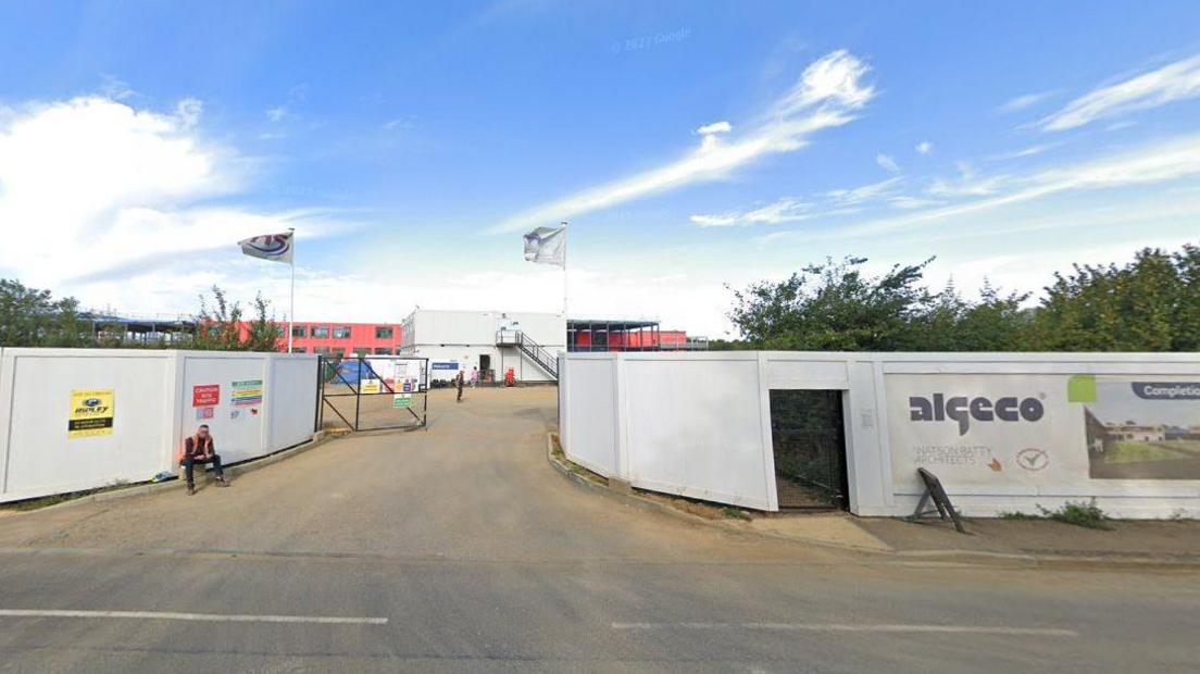 Construction site showing white boards screening the site and a two-storey red school building visible in the background. A worker in an orange hi-vis is sitting in front of the boards