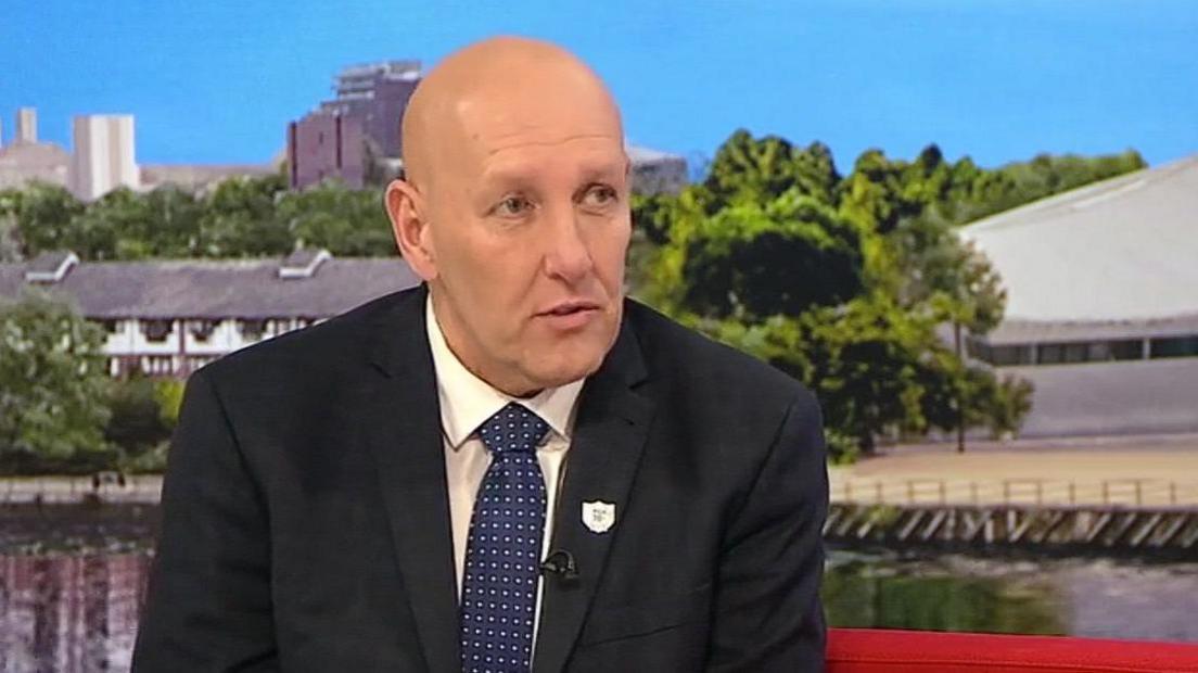 Mark Fairhurst with very short hair wearing a dark jacket with a small badge, a white shirt and a blue tie with white dots. He is sitting on a BBC red sofa with houses, trees and sky in the background.