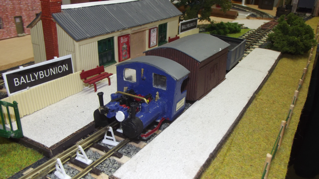 A blue engine on a monorail track is pulling a brown carriage and empty trailer. It is at Ballybunion Station which has a red bench on the platform, but is empty of passengers. There is grass and bushes to the side of the track.
