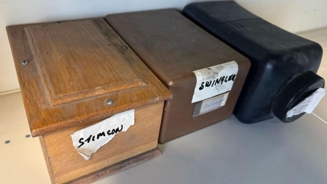 Three labelled containers on a shelf, comprising of a wooden box nailed shut labelled Stimson, a brown box labelled Swingler and a black jar on its side.