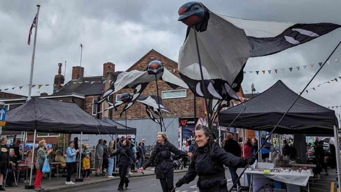 Three people in black outfits walk down a street with black market gazebos on either side. They are holding large flags in the shape of birds, which have black heads with a red spot on, and large white wings with black detailing