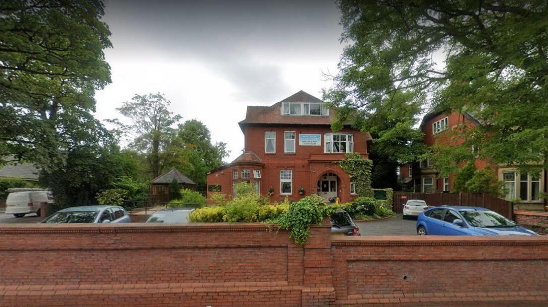 A brick wall with a red brick three storey house and a car park in front