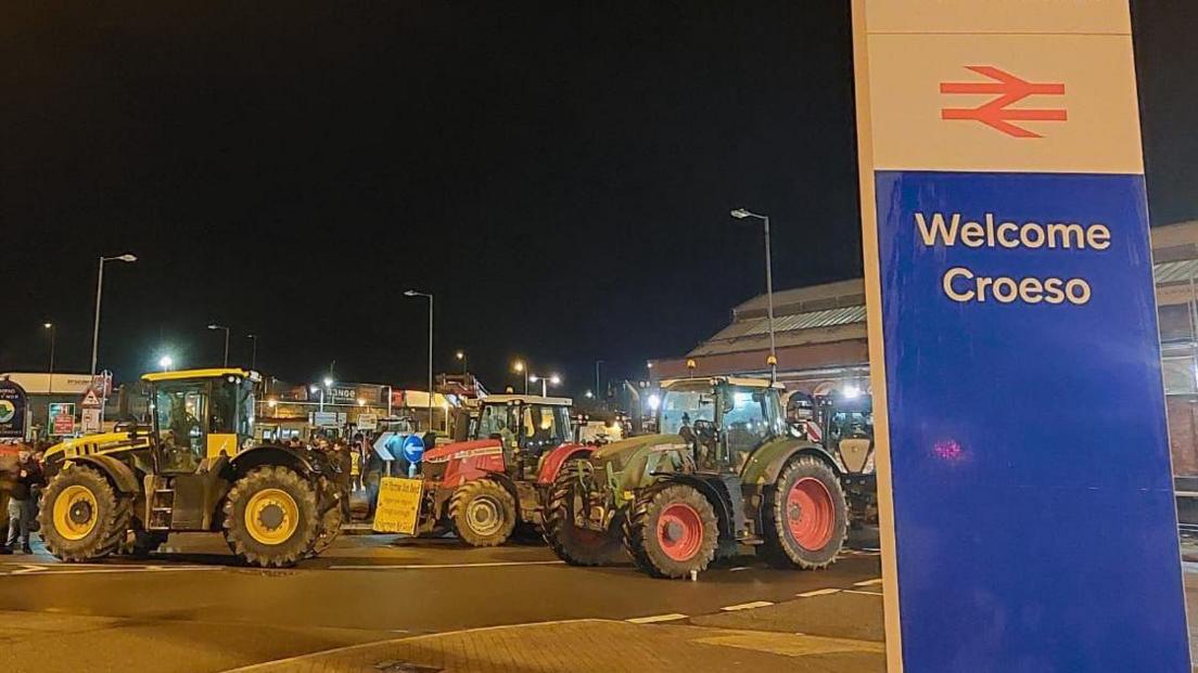 About a dozen large tractors block a road next to Holyhead port. the welcome sign with Irish Ferries and Stena Line names and logos is in the foreground