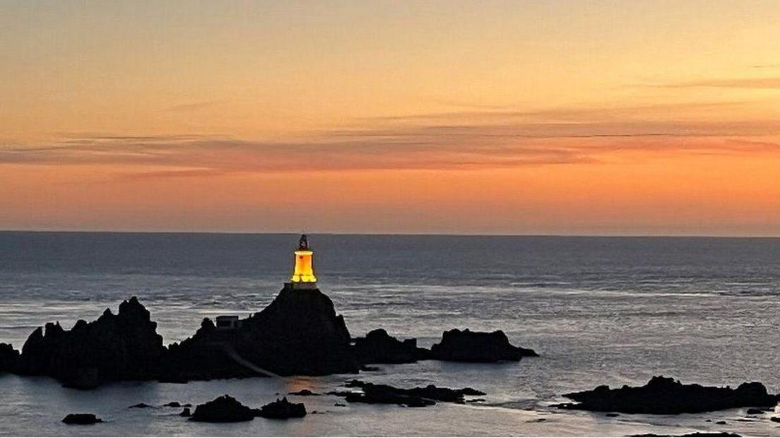 Corbière lighthouse on rocks at dusk, lit up against a sunset sky