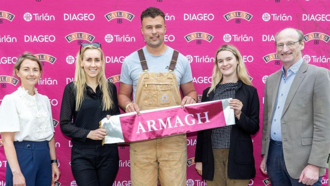 Five people standing next to each other in front of a bright pink branded background. From left to right; a woman with short brown hair, white shirt and blue trousers, then a lady with long blonde hair, black shirt and black jeans, a man with short brown hair, grey top and light brown dungarees, a woman with long blonde hair, black blazer, black and white top, and brown trousers. At the end of the row is a man who is partially bald with grey hair wearing a blue shirt, grey jacket and dark blue trousers. The three people in the middle are holding a pink banner with 'Armagh' are written on it.