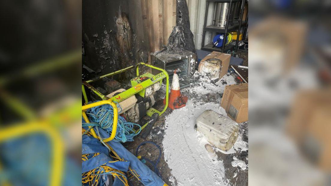 The inside of one storage container, with equipment and tools scattered about the floor. There are scorched burn marks up the metal corrugated walls and pitch-marking equipment has been melted, leaving white paint smeared all over the floor. 