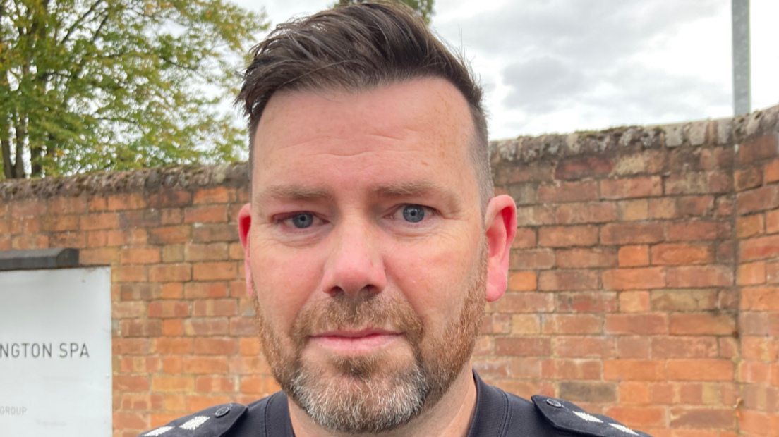 A man looks directly at the camera. He has a short beard, and his dark coloured hair is swept up and to one side.