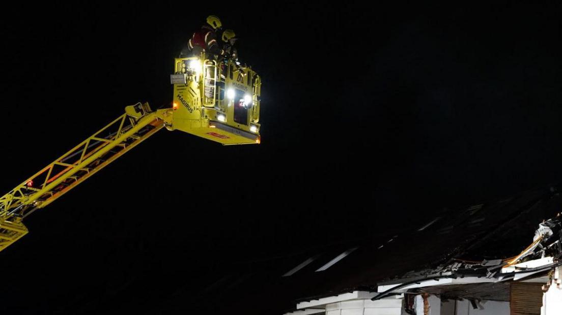 A cherry-picker with fire crew in it looms above an exploded roof