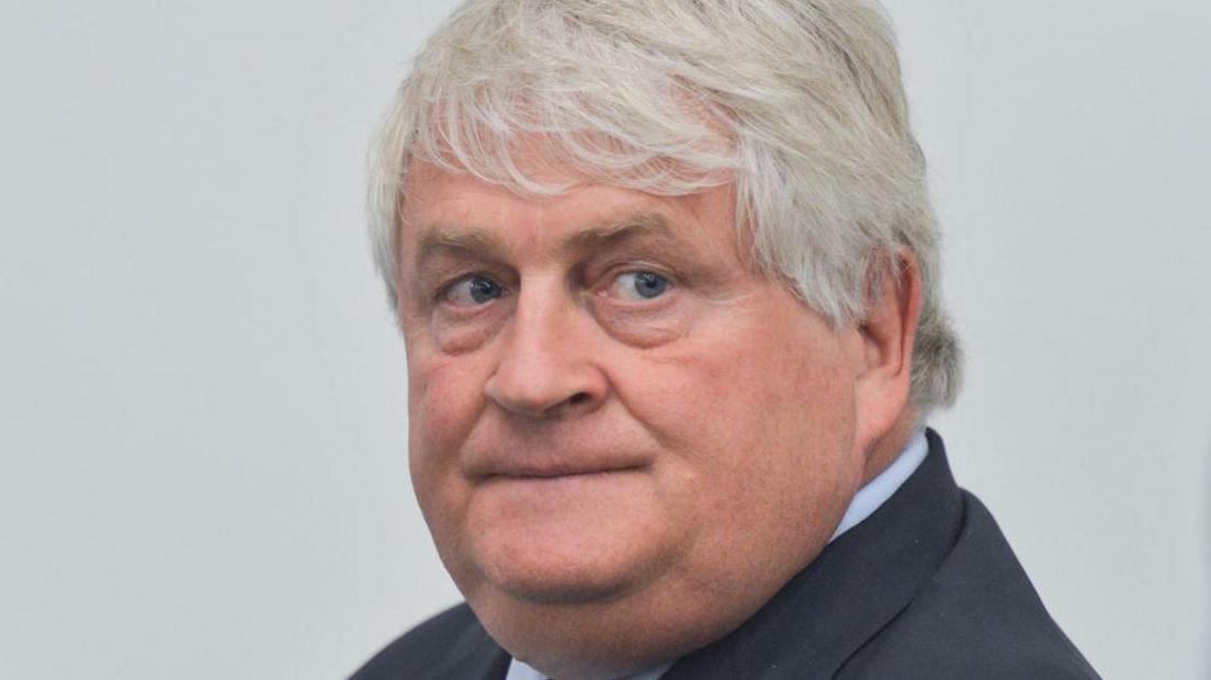 Denis O'Brien, facing the camera, with grey hair, wearing dark grey jacket and lighter shirt.